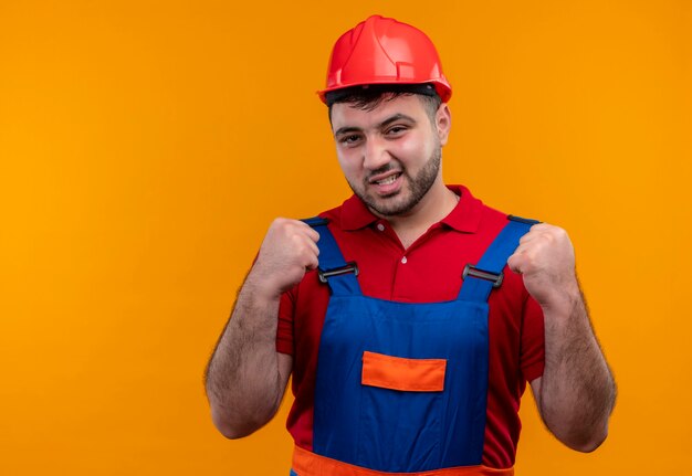 Jonge bouwer man in bouw uniform en veiligheidshelm balde vuisten blij en verlaten verheugend zijn succes