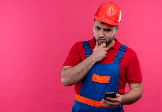 Jonge bouwer man in bouw uniform en veiligheidshelm smartphone kijken opzij met peinzende uitdrukking
