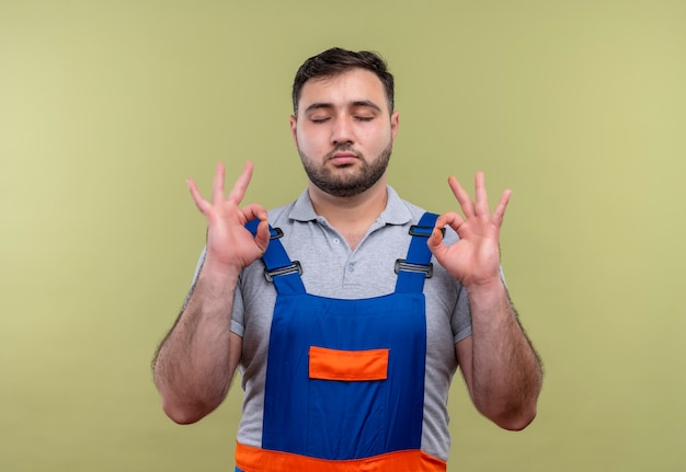 Gratis foto jonge bouwer man in bouw uniform met gesloten ogen ontspannen, meditatie gebaar met vingers maken