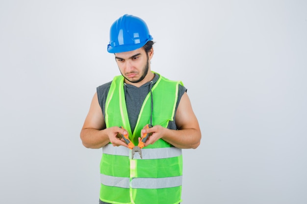 Jonge bouwer man in werkkleding uniform met tang en op zoek peinzend, vooraanzicht.