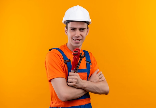 Jonge bouwer man met bouw uniform en veiligheidshelm glimlacht en houdt een groeftang