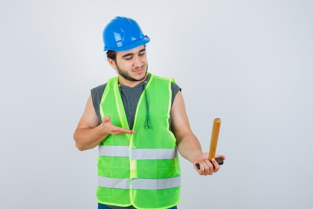 Jonge bouwer man met hamer in uniforme werkkleding en op zoek vrolijk. vooraanzicht.