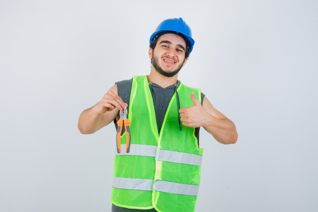 Jonge bouwer man met tang terwijl duim opdagen in uniform werkkleding en op zoek vrolijk. vooraanzicht.