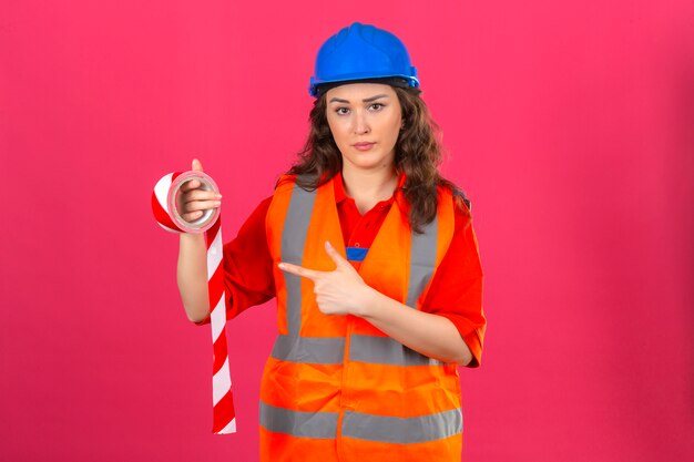 Jonge bouwer vrouw in uniform bouw en veiligheidshelm permanent met plakband en wijzend op het met wijsvinger fronsen over geïsoleerde roze muur