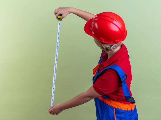 Jonge bouwer vrouw in uniform uitrekkende meetlint geïsoleerd op olijfgroene muur