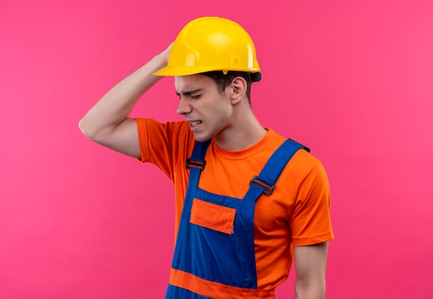 Jonge bouwersmens die bouwuniform en veiligheidshelm dragen die aan hoofdpijn lijden en zijn hoofd met de rechterhand van hoofdpijn houden