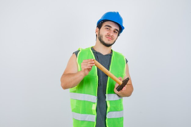 Jonge bouwersmens die hamer in uniform werkkleding toont en tevreden, vooraanzicht kijkt.