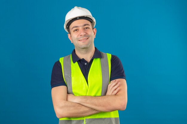 Jonge bouwersmens die witte helm en een geel vest, met het gekruiste wapens glimlachen dragen die zich op geïsoleerd blauw bevinden