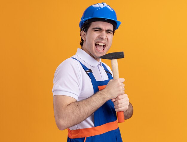 Jonge bouwersmens in bouwuniform en de hamer van de veiligheidshelm die als microfoon gebruiken die emotioneel en gelukkig status over oranje muur zingen