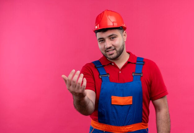 Jonge bouwersmens in bouwuniform en veiligheidshelm die kom hier gebaar met hand vriendelijk glimlachen maken