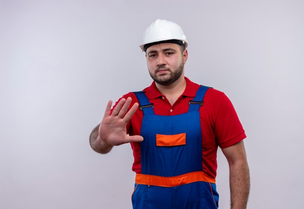 Jonge bouwersmens in bouwuniform en veiligheidshelm die met open hand stopbord met ernstig gezicht maken