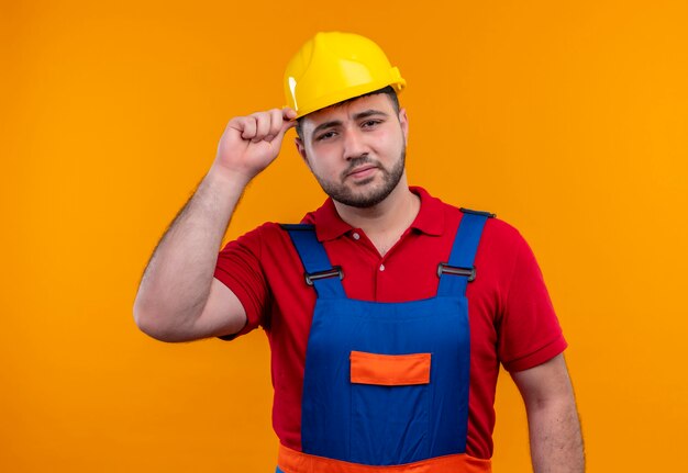 Jonge bouwersmens in bouwuniform en veiligheidshelm die opzij kijken met sceptische uitdrukking wat betreft helm
