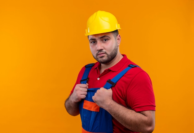 Gratis foto jonge bouwersmens in bouwuniform en veiligheidshelm hand in hand op borst die ar camera met ernstige vertrouwelijke uitdrukking kijkt