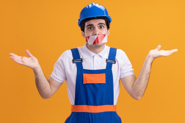 Jonge bouwersmens in bouwuniform en veiligheidshelm met tape rond de mond op zoek verward schouderophalend staande over oranje muur