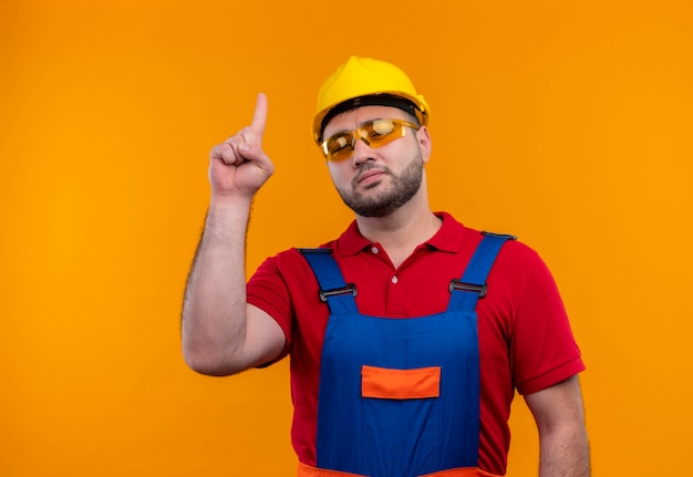 Jonge bouwersmens in bouwuniform en veiligheidshelm op zoek zelfverzekerd wijzend met wijsvinger omhoog over oranje achtergrond