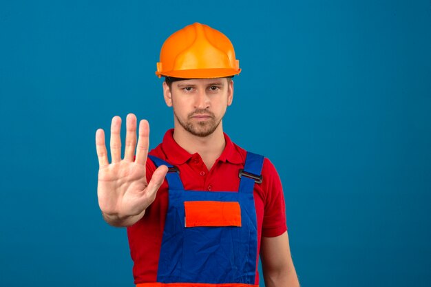 Jonge bouwersmens in eenvormige bouw en veiligheidshelm die zich met open hand bevinden die eindeteken met het ernstige en zekere gebaar van de uitdrukkingsdefensie doen over geïsoleerde blauwe muur
