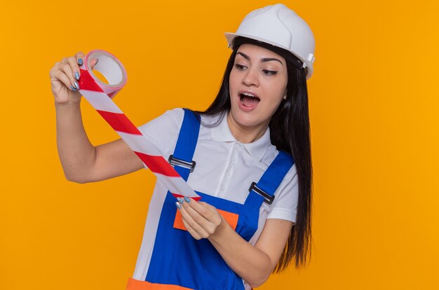 Jonge bouwersvrouw in bouwuniform en veiligheidshelm die plakband houden die het verbaasd en verbaasd zich over oranje muur bekijken