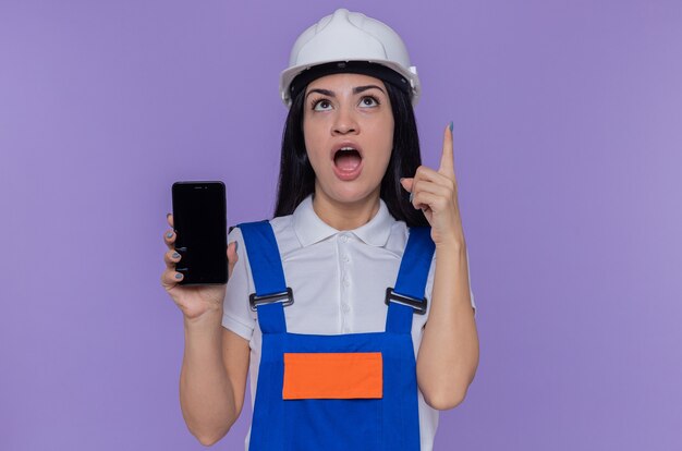 Jonge bouwersvrouw in bouwuniform en veiligheidshelm die smartphone tonen die omhoog verbaasd toont wijsvinger die geweldig idee heeft die zich over purpere muur bevindt