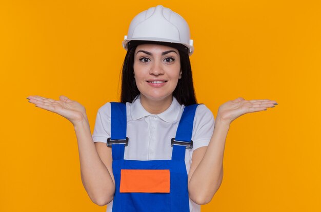 Jonge bouwersvrouw in bouwuniform en veiligheidshelm die voorzijde glimlachen die vrolijk wapens spreidt naar de zijkanten die zich over oranje muur bevinden