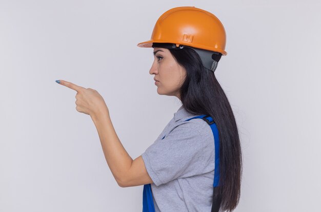 Jonge bouwersvrouw in bouwuniform en veiligheidshelm die zich zijwaarts met wijsvinger bevindt die naar iets richt dat zelfverzekerd over witte muur kijkt