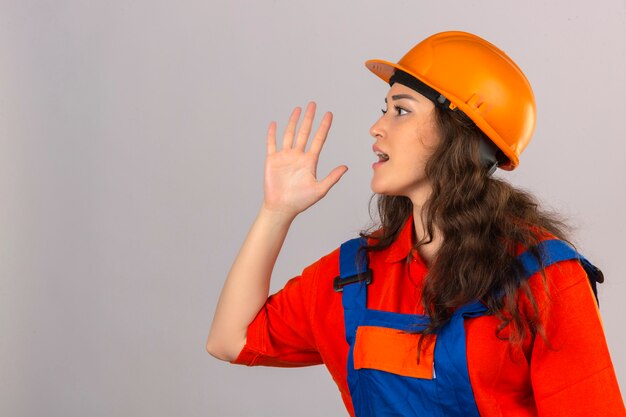 Jonge bouwersvrouw in eenvormige bouw en veiligheidshelm die aan kant met hand op mond over geïsoleerde witte muur schreeuwen