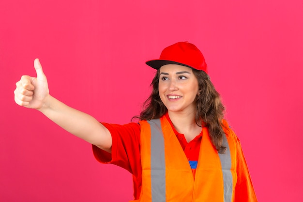 Jonge bouwersvrouw in eenvormige bouw en veiligheidshelm die duimen tonen aan iemand vriendschappelijk glimlachen over geïsoleerde roze muur