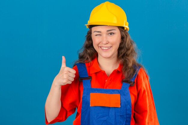 Jonge bouwersvrouw in eenvormige bouw en veiligheidshelm die het tonen van duimen met gelukkig gezicht over geïsoleerde blauwe muur knipogen