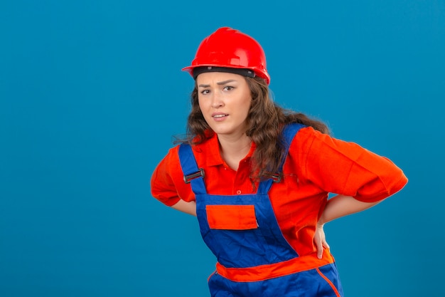 Jonge bouwersvrouw in eenvormige bouw en veiligheidshelm die ongelukkig en aan rugpijn over geïsoleerde blauwe muur kijken kijken