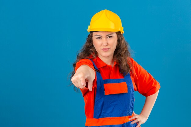 Jonge bouwersvrouw in eenvormige bouw en veiligheidshelm die ontevreden en gefrustreerd aan de camera boos en woedend met u over geïsoleerde blauwe muur richten