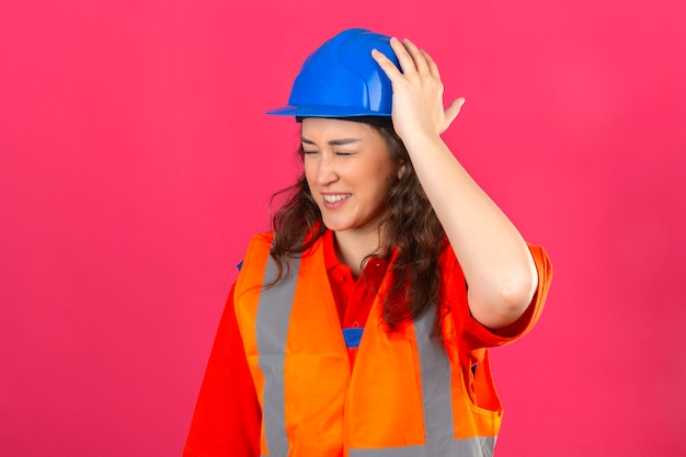 Jonge bouwersvrouw in eenvormige bouw en veiligheidshelm die onwel het raken wat betreft hoofd hebben die pijn over geïsoleerde roze muur hebben