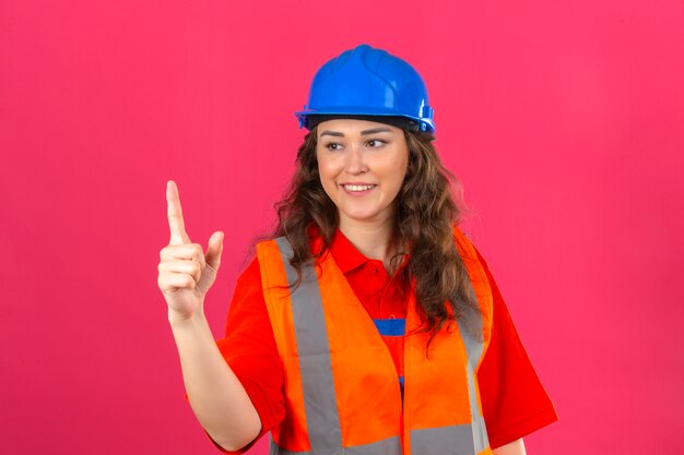 Jonge bouwersvrouw in eenvormige bouw en veiligheidshelm die tonend nummer één met vingers over geïsoleerde roze muur glimlachen