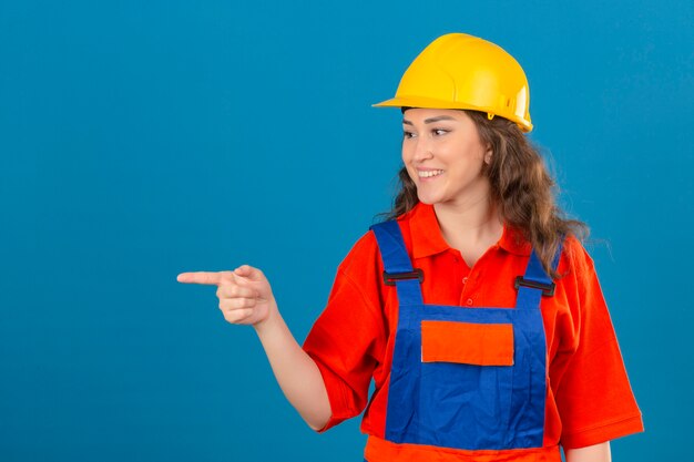 Jonge bouwersvrouw in eenvormige bouw en veiligheidshelm die vrolijk het kijken zeker het richten met wijsvinger aan de kant over geïsoleerde blauwe muur glimlachen