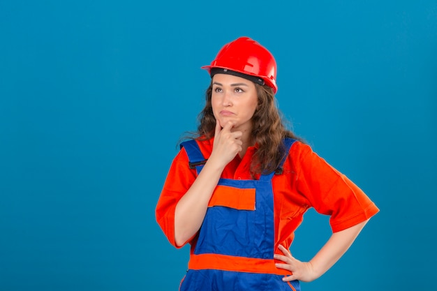 Jonge bouwersvrouw in eenvormige bouw en veiligheidshelm die zich met hand bij kin bevinden die hebbend over geïsoleerde blauwe muur denken