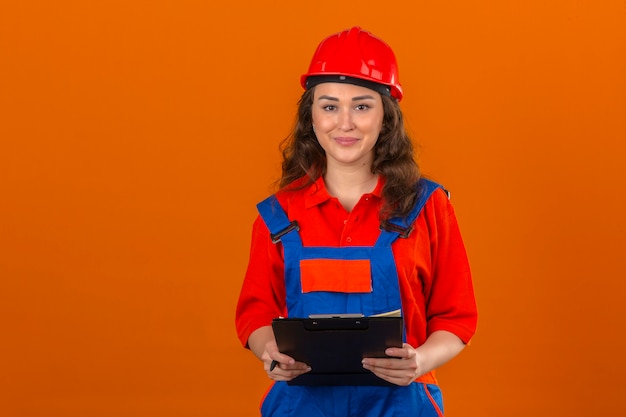 Jonge bouwersvrouw in eenvormige bouw en veiligheidshelm die zich met klembord bevinden die het zekere glimlachen vriendschappelijk over geïsoleerde oranje muur kijken