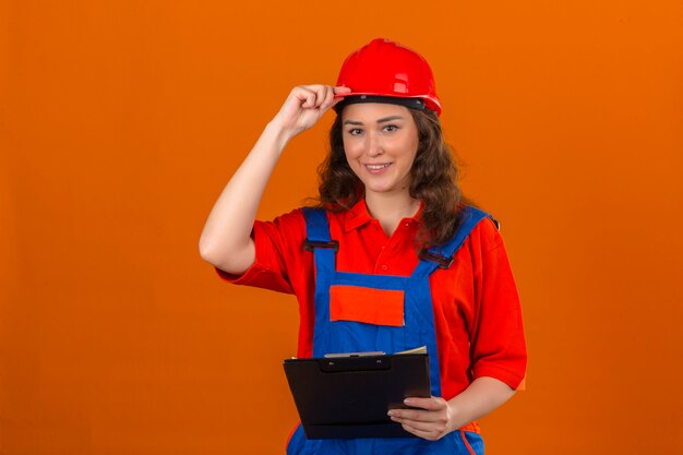 Jonge bouwersvrouw in eenvormige bouw en veiligheidshelm die zich met klembord bevinden glimlachen vriendschappelijk wat betreft haar helm over geïsoleerde oranje muur