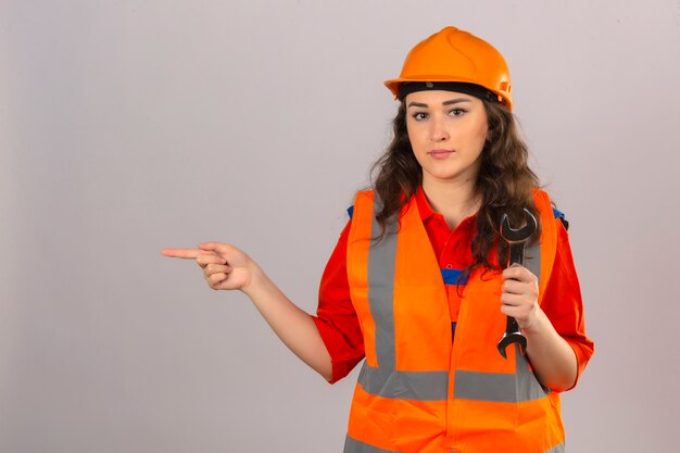 Jonge bouwersvrouw in eenvormige bouw en veiligheidshelm die zich met moersleutel bevinden die wijsvinger aan de kant over geïsoleerde witte muur richten