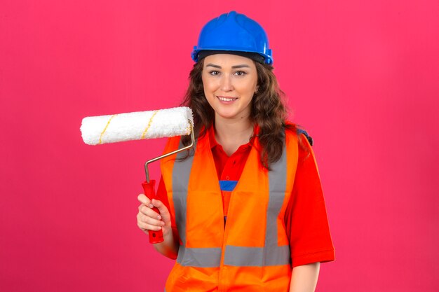 Jonge bouwersvrouw in eenvormige bouw en veiligheidshelm die zich met verfrol bevinden die met glimlach op gezicht camera over geïsoleerde roze muur bekijken