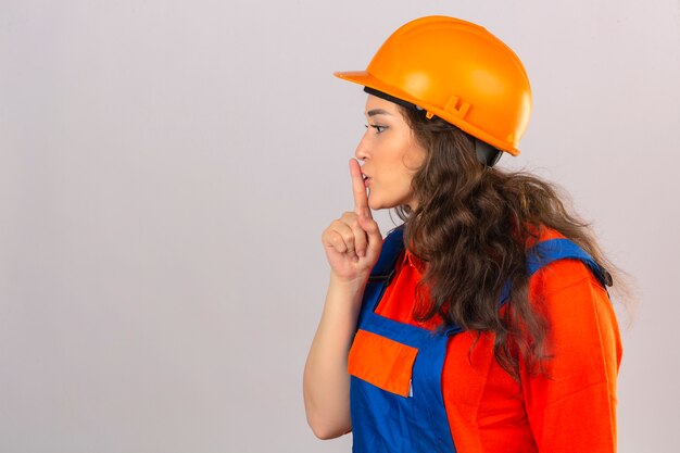 Jonge bouwersvrouw in eenvormige bouw en veiligheidshelm met haar vinger aan haar lippen of het maken van gebaar van stilte over geïsoleerde witte muur