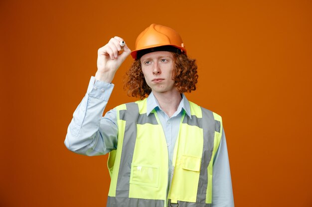 Jonge bouwvakker in bouwuniform en veiligheidshelm die een marker vasthoudt die iets in de lucht schrijft en er zelfverzekerd uitziet over een oranje achtergrond