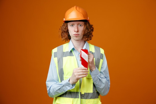 Gratis foto jonge bouwvakker in bouwuniform en veiligheidshelm met plakband die naar camera kijkt met een serieus gezicht over oranje achtergrond