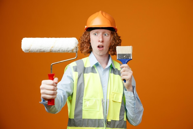 Jonge bouwvakker in bouwuniform en veiligheidshelm met verfroller en penseel die er verward uitziet en twijfels heeft over oranje achtergrond