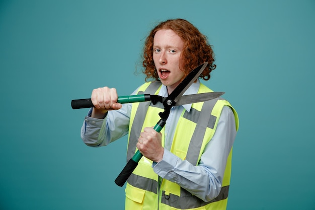 Jonge bouwvakker in bouwuniform met een schaar die naar de camera kijkt terwijl hij opgewonden en gelukkig over een blauwe achtergrond staat