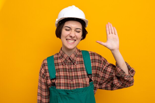 Jonge bouwvrouw in bouwuniform en veiligheidshelm blij en opgewonden glimlachend vrolijk met open palm die over oranje muur staat