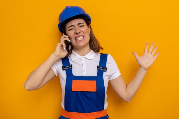 Jonge bouwvrouw in bouwuniform en veiligheidshelm die er geïrriteerd en geïrriteerd uitziet terwijl ze op een mobiele telefoon praat die op oranje staat