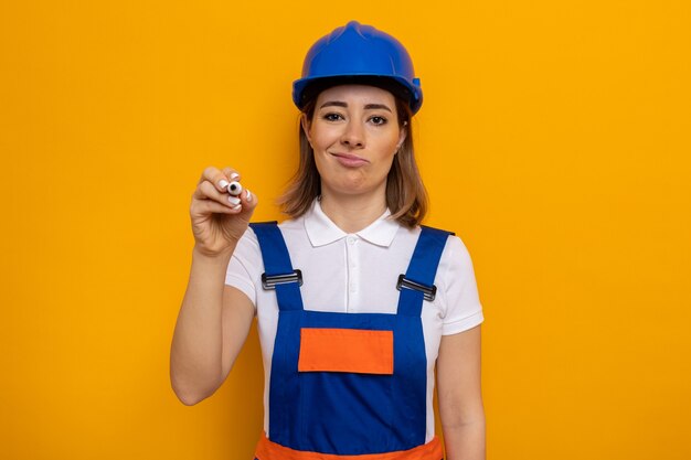 Jonge bouwvrouw in bouwuniform en veiligheidshelm die met een sceptische glimlach op het gezicht kijkt en met pen in de lucht schrijft