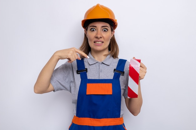 Jonge bouwvrouw in bouwuniform en veiligheidshelm met plakband die met de wijsvinger erop wijst en er verward uitziet over een witte muur