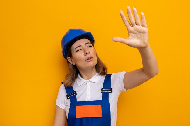 Jonge bouwvrouw in bouwuniform en veiligheidshelm opzij kijkend met loensende ogen die arm op oranje staande houden