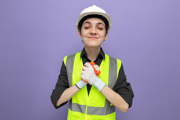 Jonge bouwvrouw in bouwvest en veiligheidshelm in rubberen handschoenen kijkend naar de voorkant glimlachend vrolijk hand in hand samen staande over paarse muur