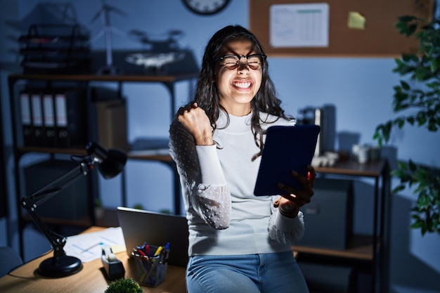 Jonge braziliaanse vrouw die 's nachts touchpad gebruikt en op kantoor werkt, erg blij en opgewonden doet winnaargebaar met opgeheven armen glimlachend en schreeuwend voor het concept van de viering van het succes