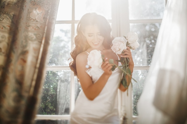 Gratis foto jonge bruid houdt een bouuqet van pioenrozen plaatsing op de vensterbank in de heldere ochtend